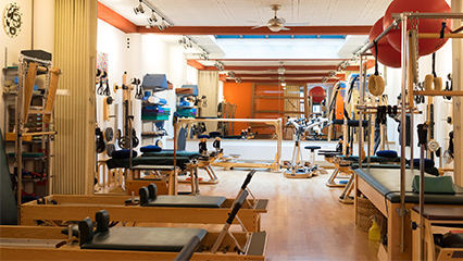 A spacious Pilates studio with wooden floors, various equipment including reformers, exercise balls, and weights, and orange and white walls. Natural light fills the room.