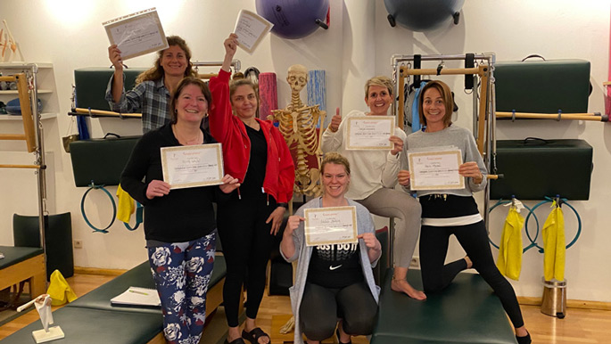 Seven people holding certificates, smiling, and posing in a pilates studio with pilates equipment and a skeleton model in the background.