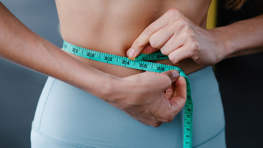 Une personne mesure sa taille à l'aide d'un ruban à mesurer sarcelle, alors qu'elle porte un pantalon de sport bleu clair.