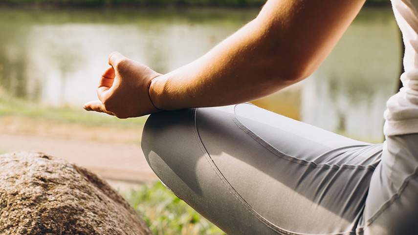 A person in a seated meditation pose outdoors beside a body of water, with one arm resting on their knee.