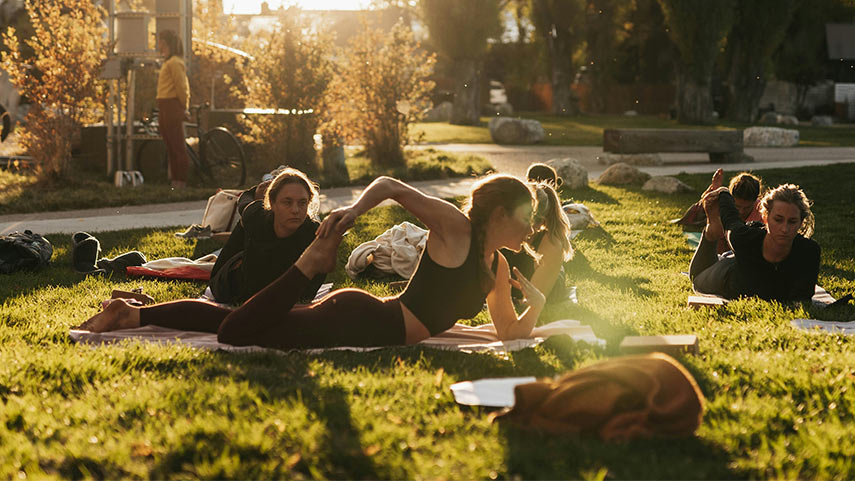 Personnes pratiquant le Pilates en plein air sur un terrain herbeux au coucher du soleil, avec des arbres et un bâtiment en arrière-plan.