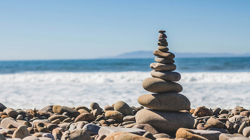 Une pile de pierres lisses est empilée en équilibre sur une plage rocheuse, avec l'océan et la terre lointaine visibles à l'arrière-plan sous un ciel clair.