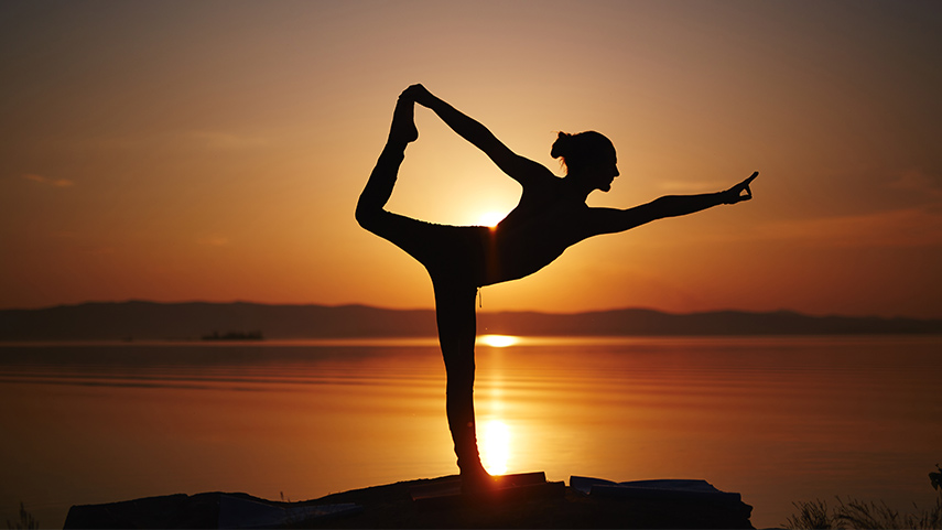A person practices a yoga pose on one leg at sunset by a calm body of water, with the sun low on the horizon and mountains in the background.