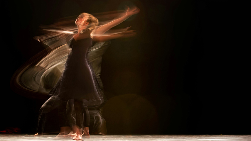 A person in a black dress dances on stage, captured with motion blur, creating sweeping light trails against a dark background.