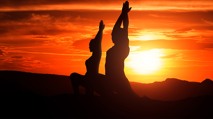 Two people practicing yoga in a kneeling pose at sunset, silhouetted against an orange sky with the sun low on the horizon and hills in the background.