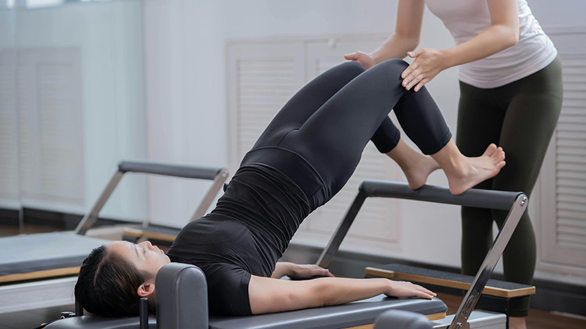 A person lying on a Pilates reformer performs a bridge exercise with support from an instructor standing nearby.
