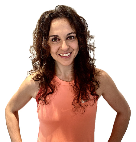 Woman with long curly brown hair wearing a sleeveless coral top, smiling and standing with hands on hips against a white background. giorgiamanno-teacher