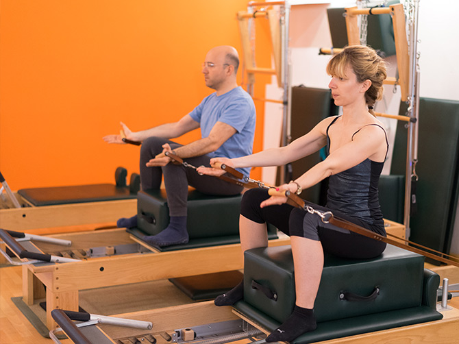 Two participants seated on Reformer boxes, extending their arms while holding the straps at Corpus Studios.